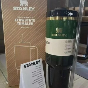 Puede incluir: Un vaso Stanley Flowstate verde y negro con pajita, con una capacidad de aproximadamente 1,18L. El vaso tiene una banda dorada y el logotipo de Stanley. La caja de al lado tiene el logotipo de Stanley y el texto "The Quencher H2.0 Flowstate Tumbler".