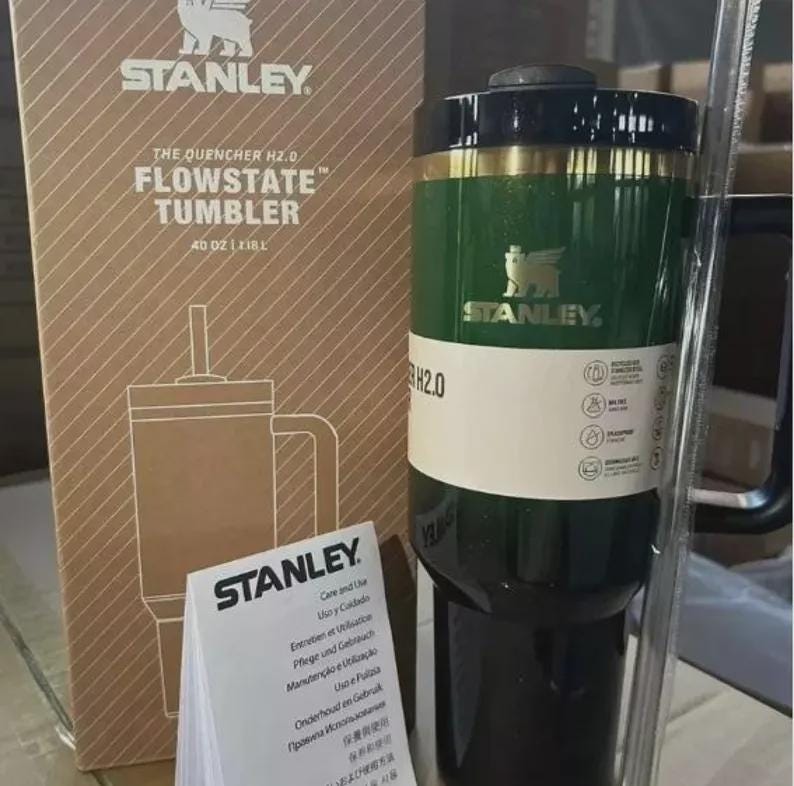 Puede incluir: Un vaso Stanley Flowstate verde y negro con pajita, con una capacidad de aproximadamente 1,18L. El vaso tiene una banda dorada y el logotipo de Stanley. La caja de al lado tiene el logotipo de Stanley y el texto "The Quencher H2.0 Flowstate Tumbler".