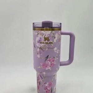 May include: A lavender Stanley travel mug with a floral pattern. The mug features a handle and a lid. The floral design includes pink and purple flowers. The brand name "STANLEY" is visible on the mug.