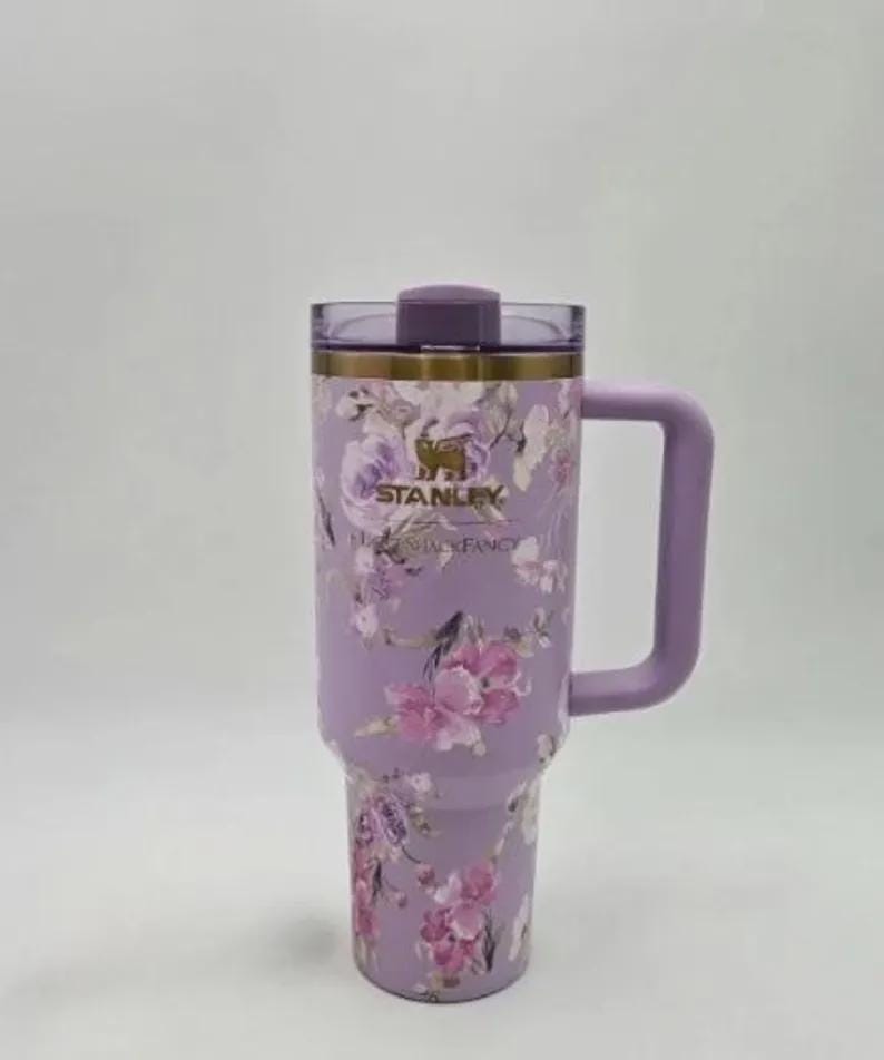 May include: A lavender Stanley travel mug with a floral pattern. The mug features a handle and a lid. The floral design includes pink and purple flowers. The brand name "STANLEY" is visible on the mug.
