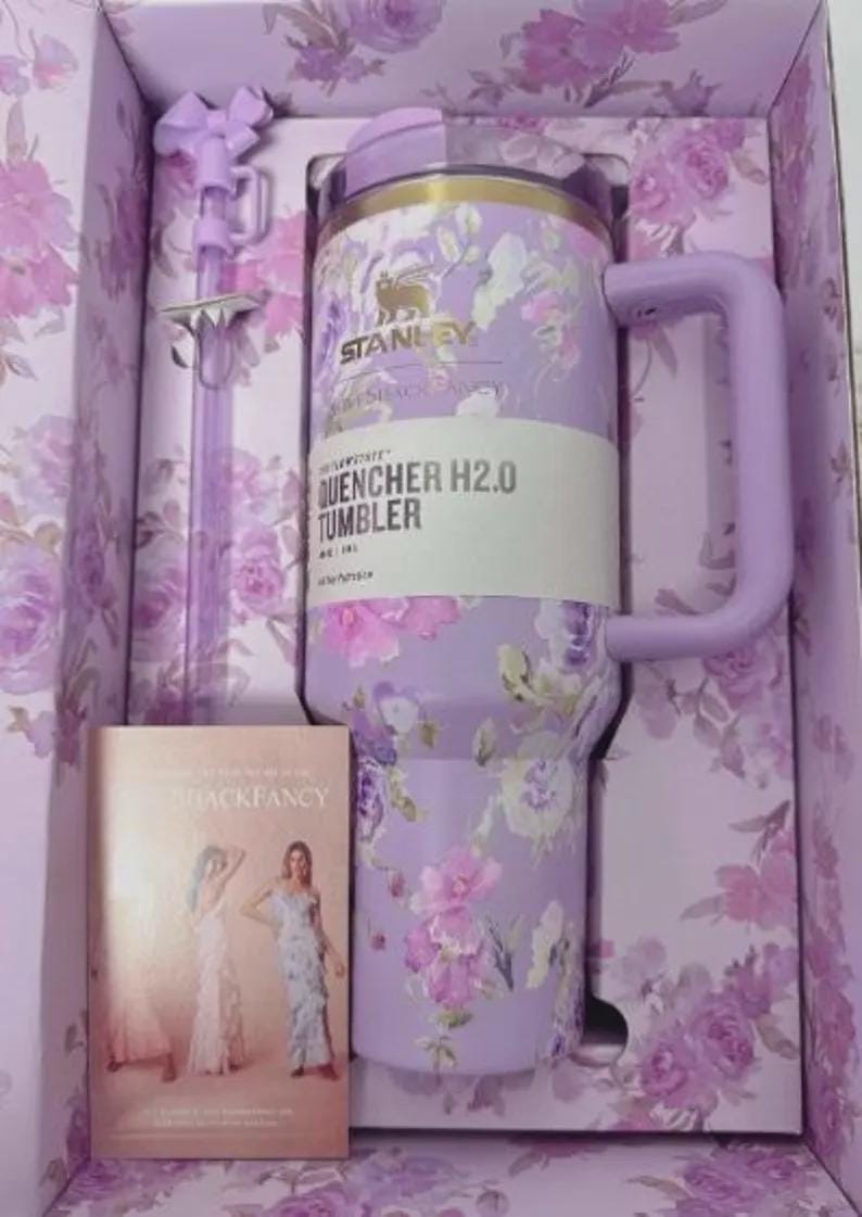 May include: A lavender Stanley Quencher H2.0 Tumbler with a floral design, handle, and straw, presented in a matching box. The tumbler features the text "QUENCHER H2.0 TUMBLER". A book with two women in dresses is also in the box.