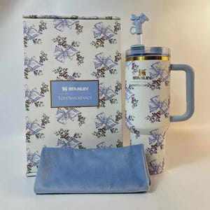 May include: A white Stanley tumbler with a blue bow and floral pattern, a matching box, and a blue cloth. The tumbler has a handle and a straw with a bow-shaped topper. The box has the text "STANLEY LOVESHACKFANCY".