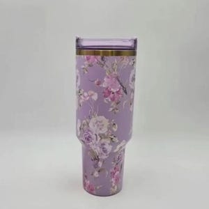 May include: A lilac-coloured tumbler adorned with a floral pattern of pink and white roses. It features a gold band at the top and a clear, rectangular lid. The design is elegant and suitable for everyday use.