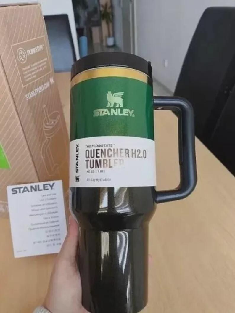 Puede incluir: Un vaso Stanley Quencher H2.0 negro y verde con asa. El vaso tiene una banda verde con el logotipo y el texto de Stanley. El producto est&aacute; dise&ntilde;ado para la hidrataci&oacute;n durante todo el d&iacute;a y tiene 1,18 litros.