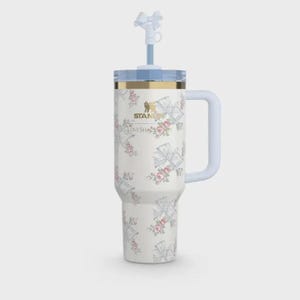May include: A white floral Stanley tumbler with a light blue handle, lid, and straw. The tumbler features a repeating pattern of bows and roses. The text "STANLEY LOVESHACK" is printed on the side. The tumbler is designed to hold approximately 30 ounces.