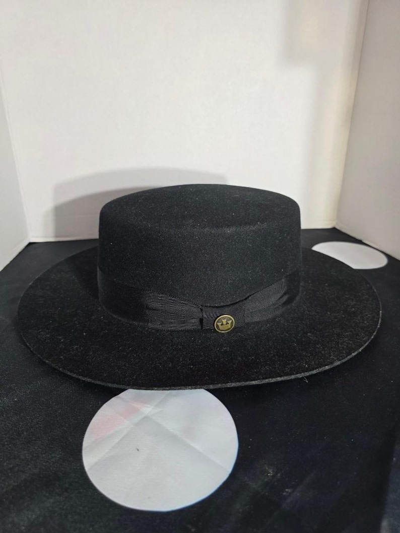 Op de afbeelding: Een zwarte hoed met een platte bovenkant en brede rand. De hoed heeft een zwarte lintband met een goudkleurig embleem. Gemaakt van een viltachtig materiaal, geschikt voor diverse gelegenheden.