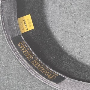 Op de afbeelding: Close-up van de binnenkant van een donkergrijze hoed met een geel label met de tekst "X-LARGE". De binnenband van de hoed heeft de gouden tekst "GOORIN BROS" en "BOLD HAT MAKERS SINCE 1895".