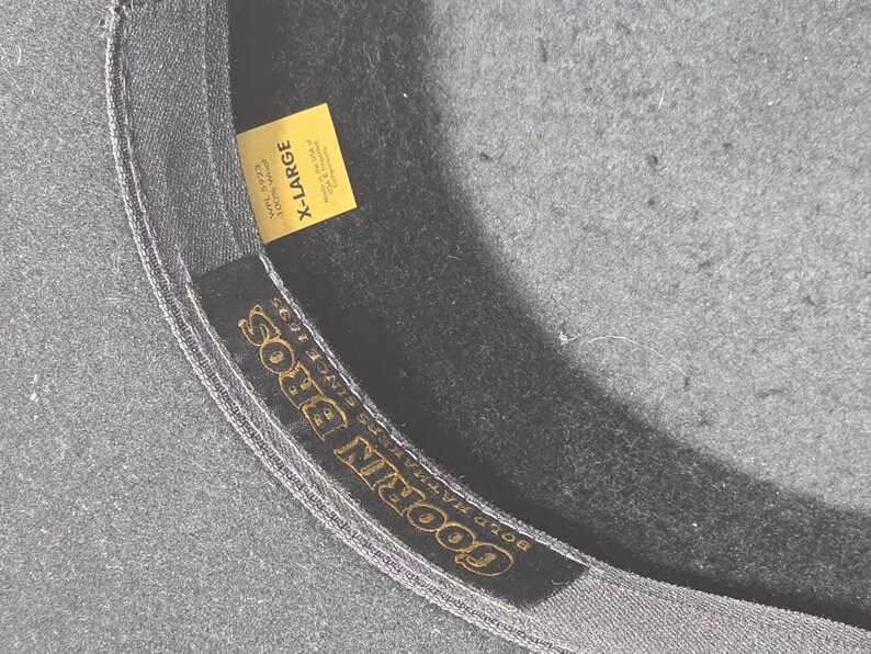 Op de afbeelding: Close-up van de binnenkant van een donkergrijze hoed met een geel label met de tekst "X-LARGE". De binnenband van de hoed heeft de gouden tekst "GOORIN BROS" en "BOLD HAT MAKERS SINCE 1895".