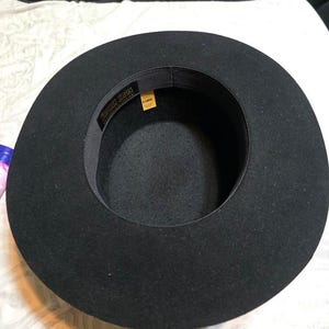 Op de afbeelding: Een zwarte fedora-achtige hoed met een brede rand. De hoed heeft een donker interieur en een band met de tekst "Fookin Bros" en een geel label met de tekst "X-LARGE". De hoed is gemaakt van een viltachtig materiaal.