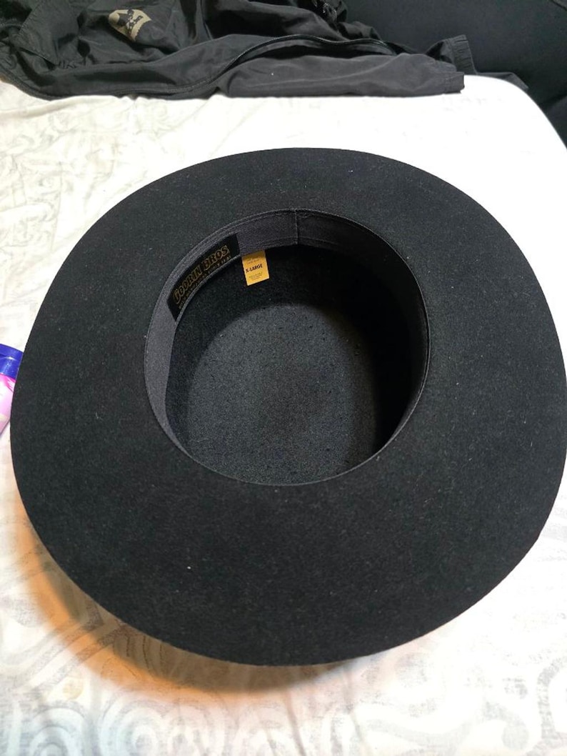 Op de afbeelding: Een zwarte fedora-achtige hoed met een brede rand. De hoed heeft een donker interieur en een band met de tekst "Fookin Bros" en een geel label met de tekst "X-LARGE". De hoed is gemaakt van een viltachtig materiaal.