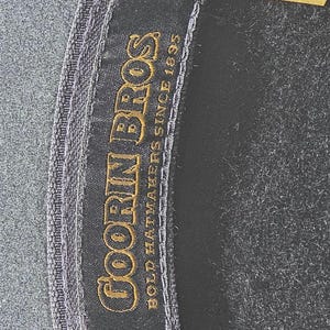 Op de afbeelding: Close-up van een donkergrijze hoed met een zwart interieur. De hoed heeft een label met de tekst "GOORIN BROS BOLD HATMAKERS SINCE 1895" in gouden letters. Een label geeft aan dat de hoed X-LARGE is en gemaakt van 100% wol.