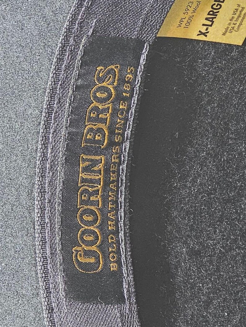 Op de afbeelding: Close-up van een donkergrijze hoed met een zwart interieur. De hoed heeft een label met de tekst "GOORIN BROS BOLD HATMAKERS SINCE 1895" in gouden letters. Een label geeft aan dat de hoed X-LARGE is en gemaakt van 100% wol.