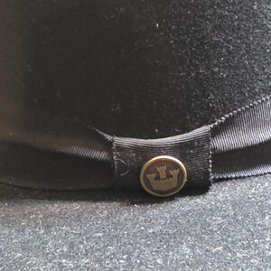 Op de afbeelding: Een zwarte fedora hoed met een zwarte lintband. De hoed heeft een goudkleurige knoop met een kroonembleem. De hoed is gemaakt van een viltachtig materiaal en is geschikt voor diverse gelegenheden.
