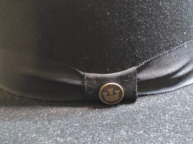 Op de afbeelding: Een zwarte fedora hoed met een zwarte lintband. De hoed heeft een goudkleurige knoop met een kroonembleem. De hoed is gemaakt van een viltachtig materiaal en is geschikt voor diverse gelegenheden.
