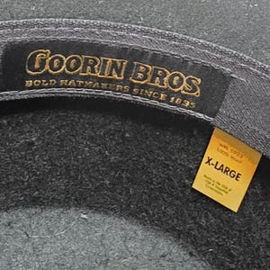 Op de afbeelding: Close-up van een donkergrijze hoed met de merknaam "Goorin Bros" in gouden letters. De hoed is gelabeld "X-LARGE" op een geel label, wat de maat aangeeft. De hoed is gemaakt van 100% wol.