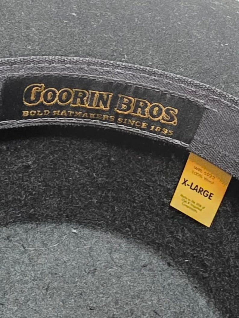 Op de afbeelding: Close-up van een donkergrijze hoed met de merknaam "Goorin Bros" in gouden letters. De hoed is gelabeld "X-LARGE" op een geel label, wat de maat aangeeft. De hoed is gemaakt van 100% wol.