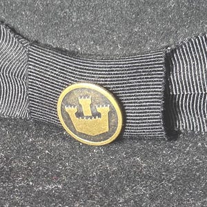 Op de afbeelding: Close-up van een donkergrijze hoed met een zwart-wit gestreept lint. Een klein, rond, goudkleurig embleem met een kasteelontwerp is gecentreerd op het lint. Het viltmateriaal van de hoed heeft een getextureerde uitstraling.