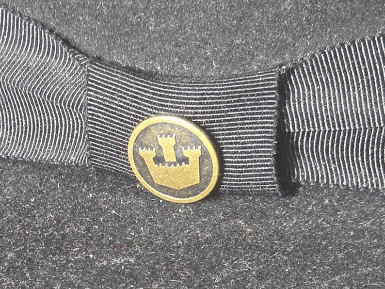 Op de afbeelding: Close-up van een donkergrijze hoed met een zwart-wit gestreept lint. Een klein, rond, goudkleurig embleem met een kasteelontwerp is gecentreerd op het lint. Het viltmateriaal van de hoed heeft een getextureerde uitstraling.