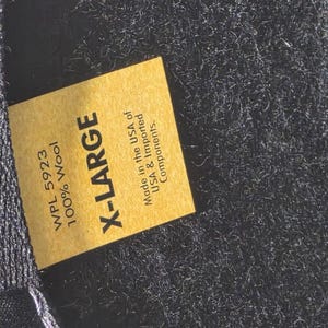 Op de afbeelding: Close-up van een gouden label met de tekst "X-LARGE" in vette zwarte letters. Het label bevat ook de tekst "WPL 5923 100% Wool" en "Made in the USA of USA & Imported Components."