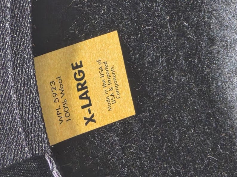 Op de afbeelding: Close-up van een gouden label met de tekst "X-LARGE" in vette zwarte letters. Het label bevat ook de tekst "WPL 5923 100% Wool" en "Made in the USA of USA & Imported Components."