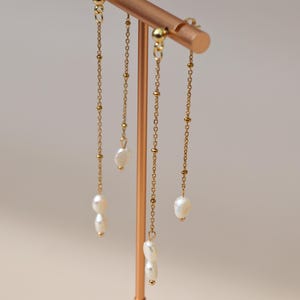 Peut inclure: Boucles d'oreilles dorées avec un design de chaîne délicat. Chaque boucle d'oreille présente une série de petites perles dorées et une seule perle blanche allongée. Les boucles d'oreilles sont présentées sur un support cuivré sur fond neutre.