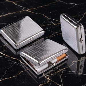May include: Three rectangular metal cigarette cases with a polished silver finish and a textured, woven-style pattern on the front. One case is open, revealing several cigarettes inside, while the other two cases are closed and positioned nearby.