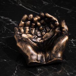 May include: A decorative bowl shaped like two cupped hands joined together. The object features a metallic bronze finish with dark, distressed black highlights, creating a textured appearance that emphasizes the contours of the fingers and palms.