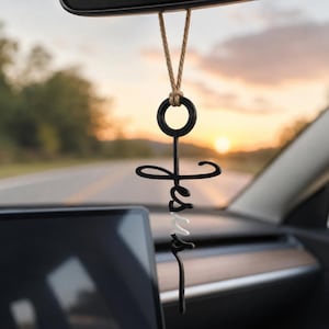 May include: A black and white hanging car ornament featuring the word Jesus in a stylized script font. The ornament is suspended by a tan twine cord and includes a circular loop at the top, forming a cross shape with the lettering.
