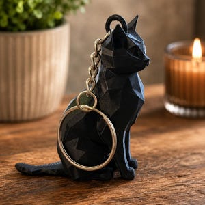 May include: A black geometric cat keyring with a gold-coloured keyring and chain. The cat figurine is sitting with its tail extended. The keyring is on a wooden surface, with a potted plant and a lit candle in the background.