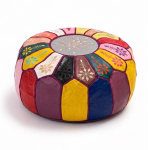 May include: A round, multi-colored leather pouf featuring a central gray circular panel with intricate white embroidery. The sides are composed of alternating vertical panels in red, yellow, blue, purple, pink, and black, accented with stitching.