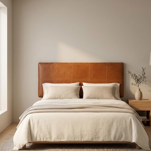 May include: A rectangular tan leather headboard with visible stitching, positioned behind a bed dressed in cream-colored linens, including two rectangular pillows and a textured throw blanket, set against a plain light-colored wall.