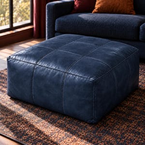 May include: A square, low-profile ottoman upholstered in dark navy blue leather. The surface features a grid-like pattern created by stitched seams, forming nine distinct rectangular sections across the top of the piece of furniture.