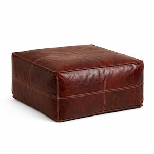 May include: A square, low-profile floor ottoman upholstered in deep brown leather with a visible, weathered texture. The surface features a cross-stitched seam pattern across the top and sides, constructed with thick, light-colored thread.
