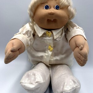 May include: A soft-sculpture doll with blonde yarn hair, blue eyes, and a single visible tooth. The doll wears a cream-colored satin collared shirt with two small yellow bows down the front and white textured fabric pants covering the feet.