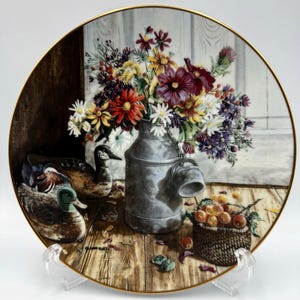 May include: A round decorative porcelain plate with a gold rim featuring a painted scene of a metal milk can filled with colorful wildflowers, two duck decoys on a wooden surface, and a small woven basket containing several small round fruits.
