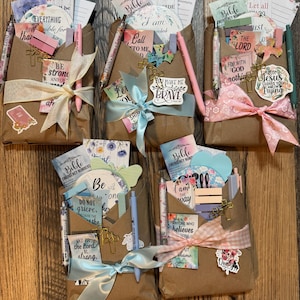 May include: Five brown paper gift envelopes containing religious stationery sets, each tied with a decorative ribbon. The sets include pens, stickers, and cards featuring floral designs and inspirational text like 'Be strong' and 'You make me brave'.