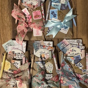 May include: Five brown paper gift bags tied with floral or solid satin ribbons, each containing assorted stationery items, pens, cross-shaped paper clips, and small cards featuring religious text and floral illustrations.