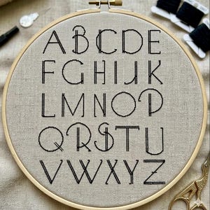May include: A completed embroidery hoop featuring the alphabet in black thread on a beige fabric. The letters are in a simple, clean font. The wooden hoop is secured with a brass screw. Additional embroidery supplies, including scissors and thread, are visible.