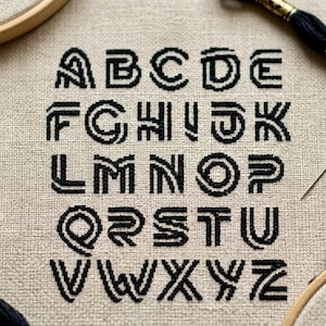 May include: Cross-stitch alphabet sampler featuring the letters A through Z in a bold, black font. The letters are stitched on a beige linen fabric, with a wooden embroidery hoop and black thread visible.