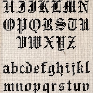 Könnte beinhalten: Ein gerahmtes Kreuzstich-Muster auf cremefarbenem Stoff, das das vollständige englische Alphabet in Groß- und Kleinbuchstaben sowie die Zahlen null bis neun zeigt, alles gestickt in einer schwarzen gotischen Schrift mit markanten Serifen.