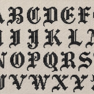 Op de afbeelding: Zwart kruissteek alfabet in gotisch lettertype, weergegeven op een crèmekleurige stof. De letters A t/m Z zijn netjes in rijen gerangschikt, wat een klassieke borduurstijl laat zien. De gedetailleerde steken creëren een textuur.