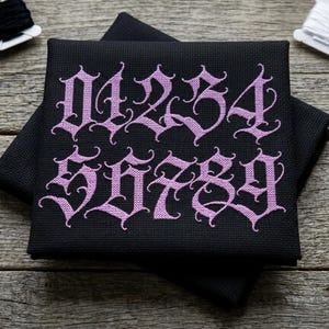 May include: Black cross-stitch fabric with the numbers 0 through 9 embroidered in a gothic font with lavender thread. The fabric is folded and resting on a wooden surface. Spools of black and white thread and small scissors are also visible.
