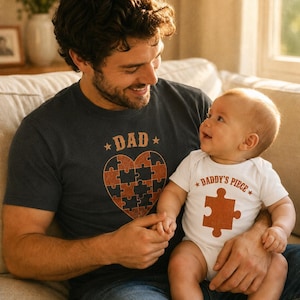 May include: A dark charcoal gray t-shirt featuring a heart-shaped puzzle graphic with the text DAD and a white baby bodysuit with a single orange puzzle piece graphic and the text DADDY'S PIECE. Both garments feature brown and orange puzzle motifs.