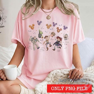 May include: A light pink t-shirt featuring a graphic print of various animated characters including Buzz Lightyear, Woody, and Jessie holding Mickey Mouse-shaped balloons. The design includes handwritten-style text labels for each character.