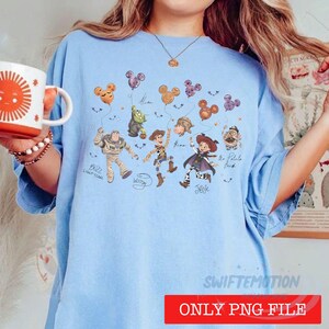 May include: A light blue t-shirt featuring a graphic print of various animated characters from the Toy Story franchise, including Buzz Lightyear, Woody, and Jessie, holding Mickey Mouse-shaped balloons, with character names written in cursive script.