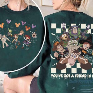 May include: A dark green crewneck sweatshirt featuring Toy Story characters in Halloween costumes. The back displays Buzz Lightyear, Woody, Jessie, and others with the text "YOU'VE GOT A FRIEND IN ME" over a checkered pattern. The front shows a smaller graphic of the characters holding Mickey-shaped balloons.