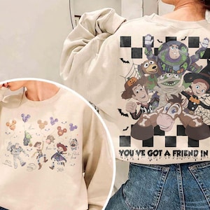May include: A beige crewneck sweatshirt featuring Toy Story characters in Halloween costumes. The back displays a large graphic of Buzz Lightyear, Woody, Jessie, and Bullseye over a black and white checkerboard pattern with the text YOU'VE GOT A FRIEND IN ME. The front shows smaller character illustrations with Mickey-shaped balloons.