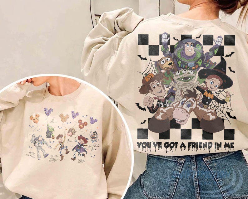 May include: A beige crewneck sweatshirt featuring Toy Story characters in Halloween costumes. The back displays a large graphic of Buzz Lightyear, Woody, Jessie, and Bullseye over a black and white checkerboard pattern with the text YOU'VE GOT A FRIEND IN ME. The front shows smaller character illustrations with Mickey-shaped balloons.
