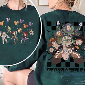 May include: A dark green crewneck sweatshirt featuring a graphic print of Toy Story characters in Halloween costumes. The design includes Buzz Lightyear, Woody, Jessie, and others with spiderwebs, bats, and the text "YOU'VE GOT A FRIEND IN ME".