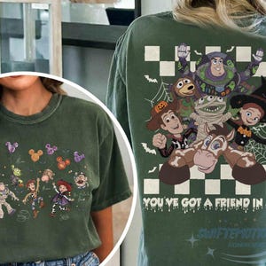 May include: A dark green t-shirt featuring Toy Story characters in Halloween costumes. The back displays Buzz Lightyear, Woody, Jessie, and others with the text "YOU'VE GOT A FRIEND IN ME". The front shows the characters holding Mickey-shaped balloons.